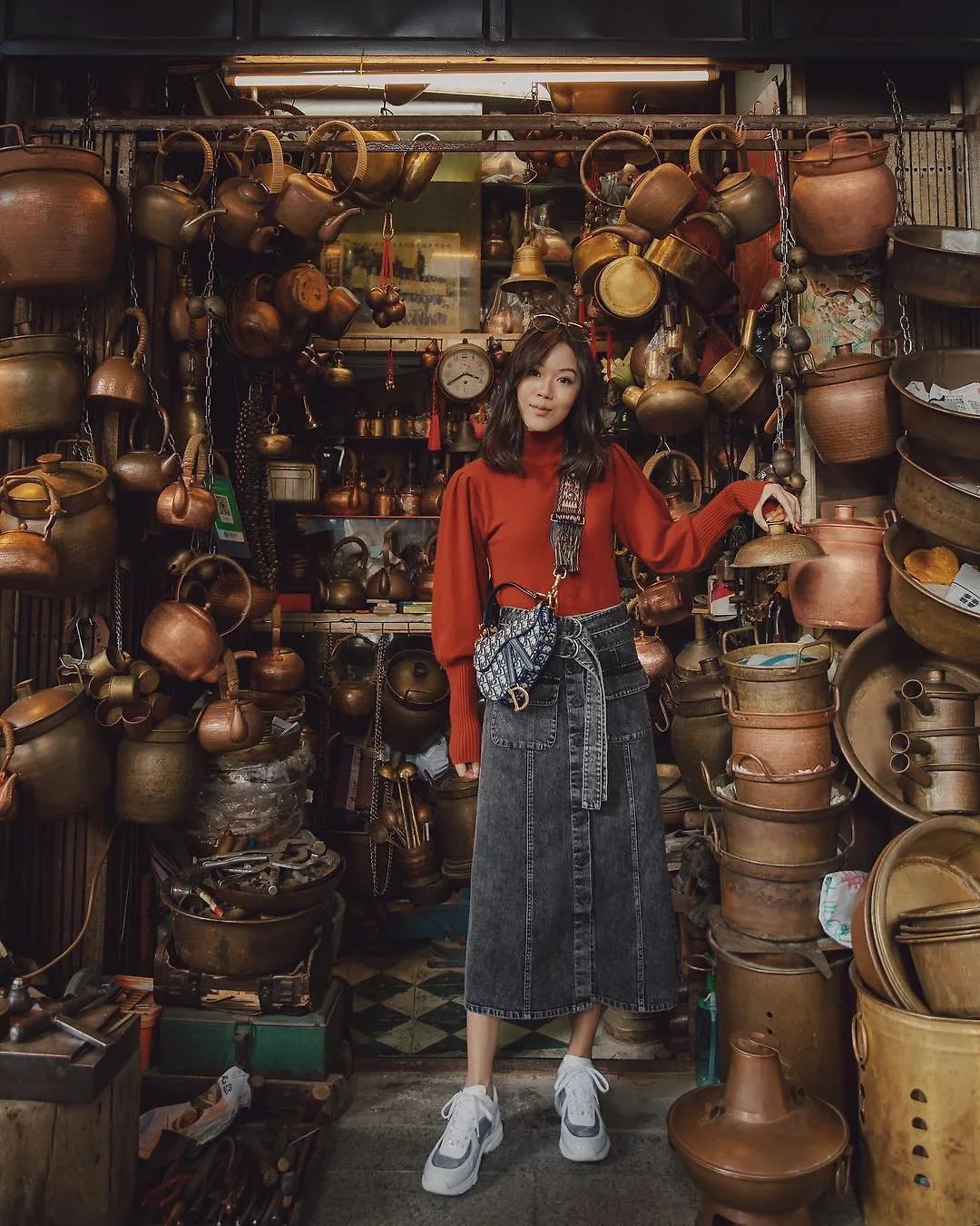 Cô gái mặc áo len đỏ và chân váy denim dài đứng trước cửa hàng đồ đồng nấu cổ điển
